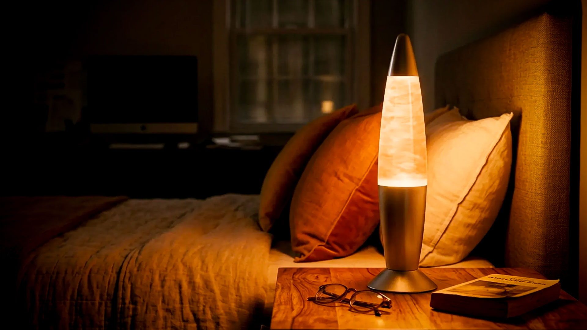 Lava lamp glows on a wooden bedside table beside pillows on a dimly lit bed in a cozy bedroom.